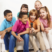 Sechs Kinder sitzen auf einem Sofa um ein Handmikrofon herum und singen.
